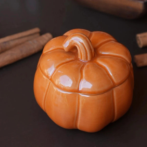 A glossy ceramic pumpkin-shaped container with a lid and a curved stem, displayed on a dark surface with cinnamon sticks and a wooden bowl in the background.