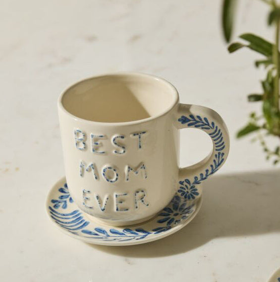 Ceramic mug with 'Best Mom Ever' text on a saucer, placed on a light surface with a plant in the background.