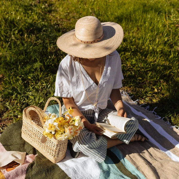Person sitting on a colorful blanket with a straw hat, picnic basket and flowers in a grassy area
