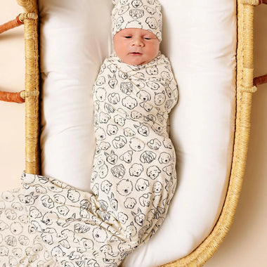 Newborn baby swaddled in a dog patterned blanket and wearing a matching hat, lying in a wicker crib.