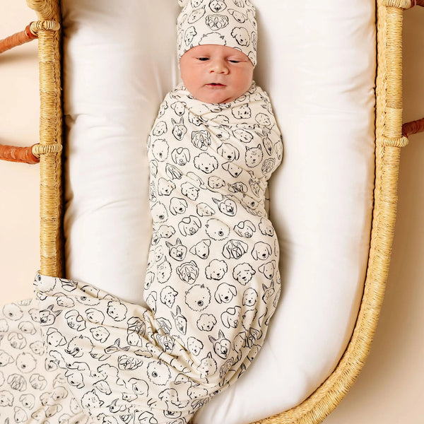Newborn baby swaddled in a dog patterned blanket and wearing a matching hat, lying in a wicker crib.