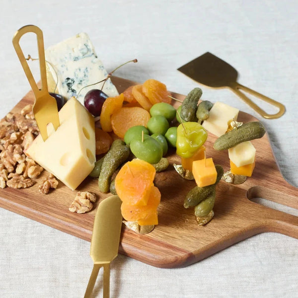 A styled charcuterie board with cheese, grapes, pickles, nuts, and skewered fruit on a wooden board, set on a light textured fabric background. Gold-toned serving utensils are arranged around the board.